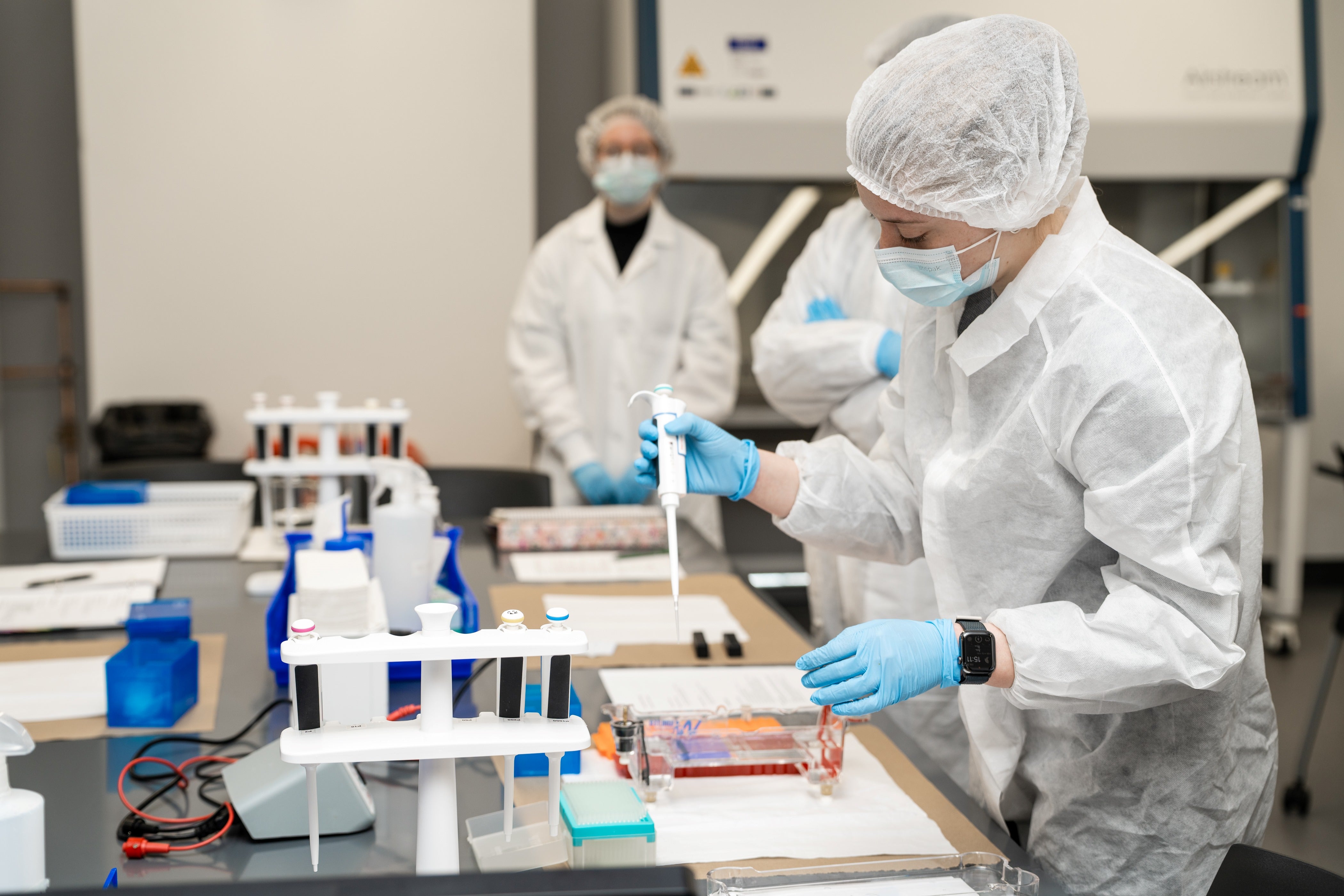 A student wearing PPE works in a biomedical lab and holds a syringe, while two others look on